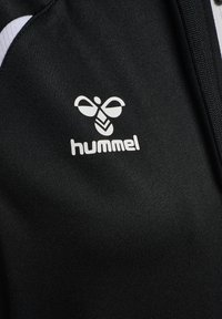 Tissu noir avec logo blanc hummel présentant une abeille stylisée au-dessus du nom de la marque sur un vêtement de sport.