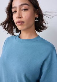 Bluza bleu deschis cu guler rotund și croială lejeră. Țesătura pare moale, cu o textură fină și detalii minime de cusături.