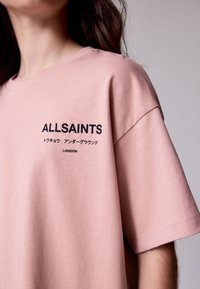 Hele roosa puuvillane T-särk lühikeste varrukatega, millel on musta tekstiga brändinimi "ALLSAINTS" ja "LONDON". Sile tekstuur.