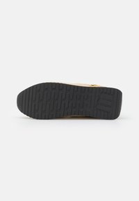 Semelle en caoutchouc noir de la basket beige et jaune, avec un motif de bande de roulement texturé et un logo ovale embossé près du talon.