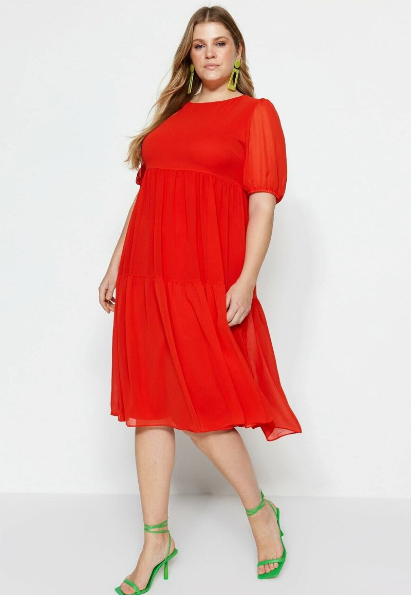 Robe rouge mi-longue avec un col rond, des manches courtes bouffantes et une jupe à volants. Portée avec des talons verts à lanières et des boucles d'oreilles remarquables.