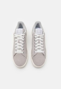 Sneakers in suede grigio con lacci e dettagli bianchi. Presentano una suola in gomma e un'etichetta con il logo sulla linguetta. Texture liscia, design low-top.