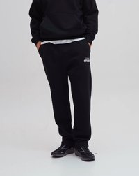 Svart sweatshirt och matchande sweatpants i mjukt material, med en logo på vänster lår och elastiska mudd. Bärs med svarta skor.