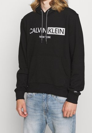 Hoodie noir en coton avec une poche avant, arborant un logo blanc "CALVIN KLEIN NEW YORK" et des cordons de serrage avec des rayures noir et blanc.