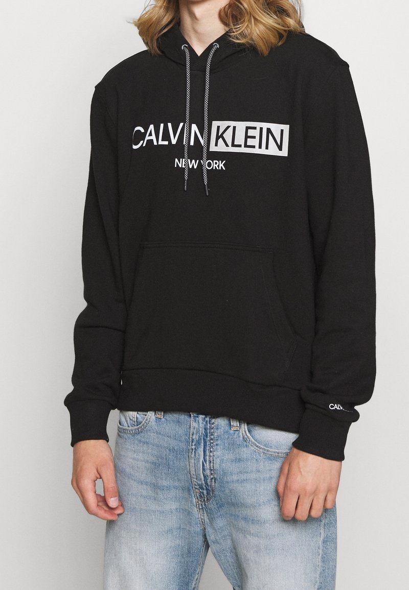 Felpa nera in cotone con tasca frontale, caratterizzata da un logo bianco "CALVIN KLEIN NEW YORK" e cordini con strisce nere e bianche.