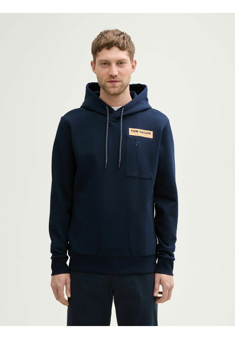 Sweat à capuche bleu marine en tissu doux avec une poche frontale et des cordons de serrage. Présente un écusson logo sur la poitrine et des poignets et ourlet côtelés.