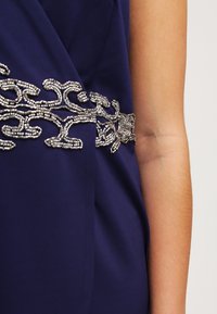 Námořnické šaty s dekorativním stříbrným korálkovým designem v pase, hladkou texturou a přiléhavým střihem.