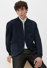 Granatowa kurtka bombowiec z gładkiego materiału z zamkiem z przodu, prążkowanym kołnierzem i mankietami oraz bocznymi kieszeniami, zestawiona z białą koszulą z guzikami.