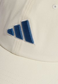 Gorra blanca con una tela texturizada que presenta un logo bordado azul con tres franjas diagonales. Costuras visibles a lo largo de los bordes.