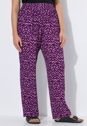 Donna con pantaloni larghi viola con motivo geometrico rosa, top nero e sandali neri open toe, in piedi su sfondo bianco.