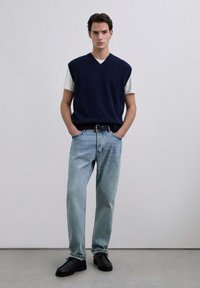 Chaleco de punto marinero sobre una camiseta blanca, jeans de mezclilla azul claro y zapatos negros. El chaleco tiene un escote en V y detalles acanalados.