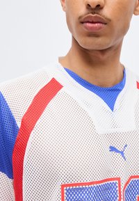 Νεαρό άτομο που φοράει λευκό αθλητικό μπλουζάκι από δίχτυ με κόκκινες και μπλε λεπτομέρειες και λογότυπο Puma στο στήθος, με φόντο μονόχρωμο.