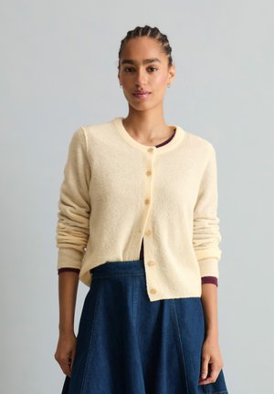 Femme aux cheveux tressés portant un cardigan crème boutonné par-dessus un haut foncé et une jupe en jean bleue, debout devant un fond clair uni.