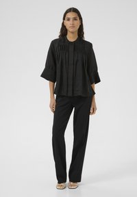 Black blouse with a round neckline, pleated design, and wide sleeves, paired with black trousers and flat sandals. Smooth texture.