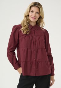 Blusa de botones color marrón con mangas largas, presenta detalle de hombros fruncidos, diseño en capas y tela texturizada. Se llevan pantalones negros.