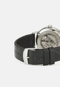 Black leather watch strap with silver stainless steel buckle. Back shows mechanical movement, glass case with branding and design details.