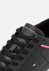 Schwarze Ledersneaker mit flachen schwarzen Schnürsenkeln, versehen mit einem Logo auf der Zunge und rot, weiß und blauen Streifen an der Seite. Sichtbare strukturierte Details.