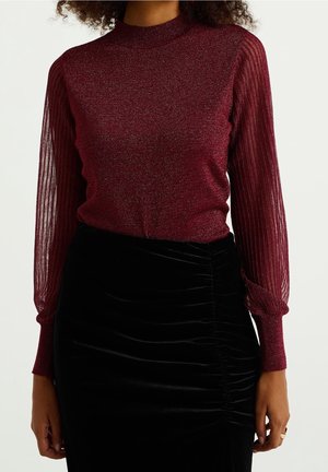 Femme portant un haut en tricot métallique bordeaux avec des manches longues rayées transparentes et un col montant, associé à une jupe noire froncée.
