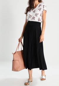 Blusa bianca floreale con maniche corte, abbinata a una gonna plissettata nera. Gli accessori includono una borsa a tracolla rosa e sandali con zeppa argentati.