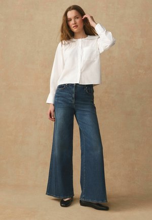 Frau mit langen Haaren, die ein weißes, geknöpftes Hemd, weit geschnittene blaue Jeans und schwarze Schuhe trägt und vor einem beigen Hintergrund posiert.