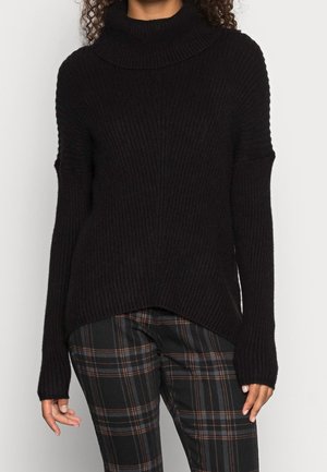 Sort ribstrikket rullekravesweater båret med mørke ternede bukser på en person med krøllet hår, stående mod en ensfarvet baggrund.