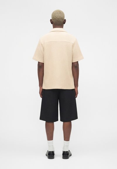 Άνδρας με κοντά ξανθά μαλλιά, φοράει μπεζ κοντομάνικο πουκάμισο, μαύρα σορτς μέχρι το γόνατο, λευκές κάλτσες και μαύρα παπούτσια, στέκεται με την πλάτη γυρισμένη.