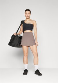Črn crop top z enim ramenom, kombiniran s rjavim krilom v stilu tenisa. Model drži črno športno torbo, obut je v črne superge in bele nogavice.
