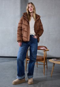 Giacca a vento marrone con cappuccio, maglione beige e jeans blu larghi. Scarpe con plateau color sabbia. Ambiente interno con una sedia e un tavolo in legno.