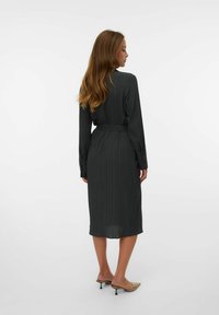 Robe longue à manches longues, de couleur gris foncé, avec une ceinture, texture côtelée et coupée au genou. Portée avec des mules à bouts ouverts à motifs clairs.