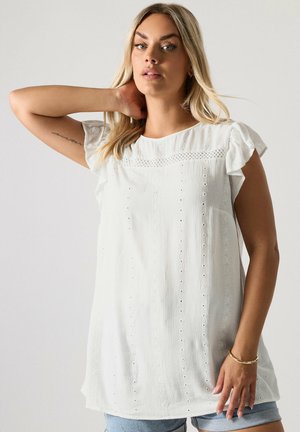 Blonde vrouw die een witte mouwloze blouse met opengewerkte details en lichtblauwe denimshorts draagt, poseert met één arm omhoog achter haar hoofd.
