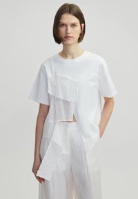 White short-sleeve T-shirt featuring layered textured panels in varying shades of white, with a relaxed fit and crew neckline.