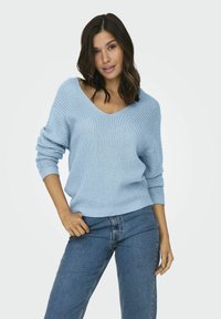 Šviesiai mėlynas megztinis su plačiu V formos kaklu, su raukšlėmis ant rankovių ir laisvu siluetu, derinamas su mėlynomis džinsomis. Minkšta tekstūra.