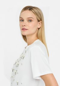 Top bianco a maniche corte con decorazioni di perle e cristalli sul davanti, tessuto liscio e scollatura tonda.