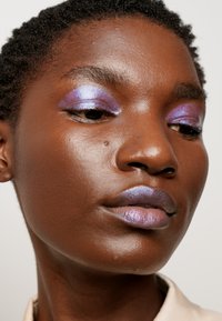Gros plan sur un visage avec des ombres à paupières violettes brillantes et un rouge à lèvres lavande irisé sur une peau noire lisse. Cheveux naturels coupés courts.