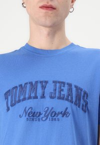 Mėlynas medvilnės marškinėliai su raukinėliu apykakle ir iškilniu tekstu: „TOMMY JEANS“ ir „New York“ tamsiai mėlyna spalva, su vintage stiliumi nuo 1985 metų.