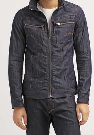 Mand iført mørk denimjakke med lynlås foran og lynlåslommer på brystet, kombineret med matchende mørke denimbukser.