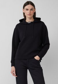 Felpa nera con cappuccio realizzata in misto cotone, dotata di una tasca frontale, chiusura con cordoncino e polsini a coste. Abbinata a pantaloni scuri.