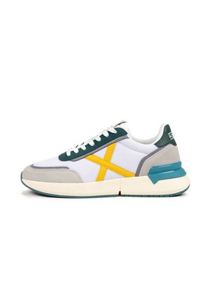 Zapatilla deportiva blanca de malla con detalles en ante gris. Cuenta con una franja diagonal amarilla y suela de goma en color verde azulado. Diseño clásico con cordones.