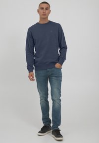Felpa navy con collo a giro, polsini e orlo a costine; abbinata a jeans blu scoloriti e sneakers nere, in piedi su uno sfondo neutro.