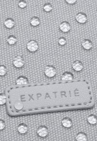Grau gefärbter Stoff mit erhabenen klaren Tropfen und einer subtilen Textur. Ein graues Lederetikett mit geprägtem Text "EXPATRIÉ" ist sichtbar.