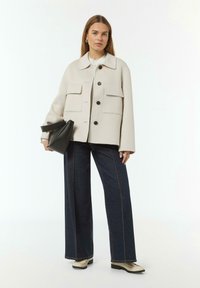 Lichtbeige oversized jas met twee voorzakken en zwarte knopen, gecombineerd met donkere wijde jeans en schoenen in goudtint. Zwarte tas.