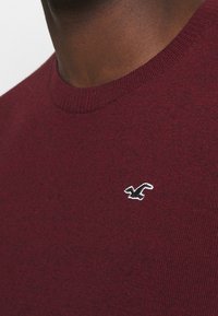 Bordeauxfärgad tröja med rund hals, ribbad krage och ett litet svart-vitt logopatch på vänster bröst. Mjuk stickad textur.