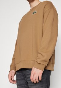 Hombre vestido con un sudadero marrón camello con un logo de cocodrilo verde en el pecho, combinado con unos jeans gris oscuro, de pie contra un fondo liso.