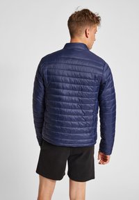 Mörkblå puffjacka med horisontellt kviltade mönster, dragkedja framtill och krage. Lättviktig, glänsande material med en avslappnad passform.