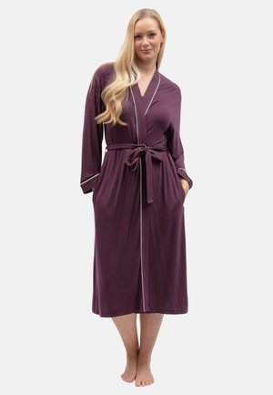 Femme aux longs cheveux blonds portant une robe violette jusqu'aux genoux avec des bordures blanches et une ceinture nouée, debout pieds nus les mains dans les poches.