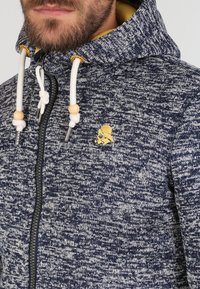 Mörkblå huvtröja med en texturerad stickad design. Har dragkedja, justerbara snören med träpärlor och en gul logotypdetalj.