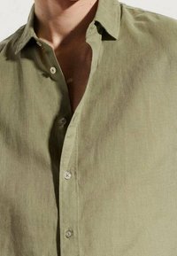 Camisa de manga corta verde oliva hecha de una tela ligera; presenta un cuello abotonado y una tapeta con botones. Textura suave.