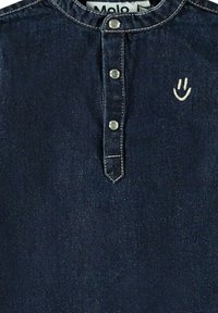 Denim-paita, jossa on pyöreä kaula-aukko, nepparivetoketju ja vaaleaa ommelta. Rintataskussa pieni valkoinen hymyilevä naama brodeerattuna.