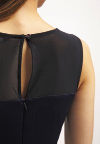 Zwarte jurk met een doorzichtig mesh bovenstuk en een nauwsluitend onderste deel. Voorzien van een knoopsluiting aan de achterkant en een klein uitsnijdetail.