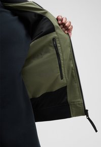 Khaki groene jacket met zwarte gaasdetails, voorzien van een ritszak aan de binnenzijde en bedrukte verzorgingsinstructies. Gladde textuur, gestructureerd ontwerp.